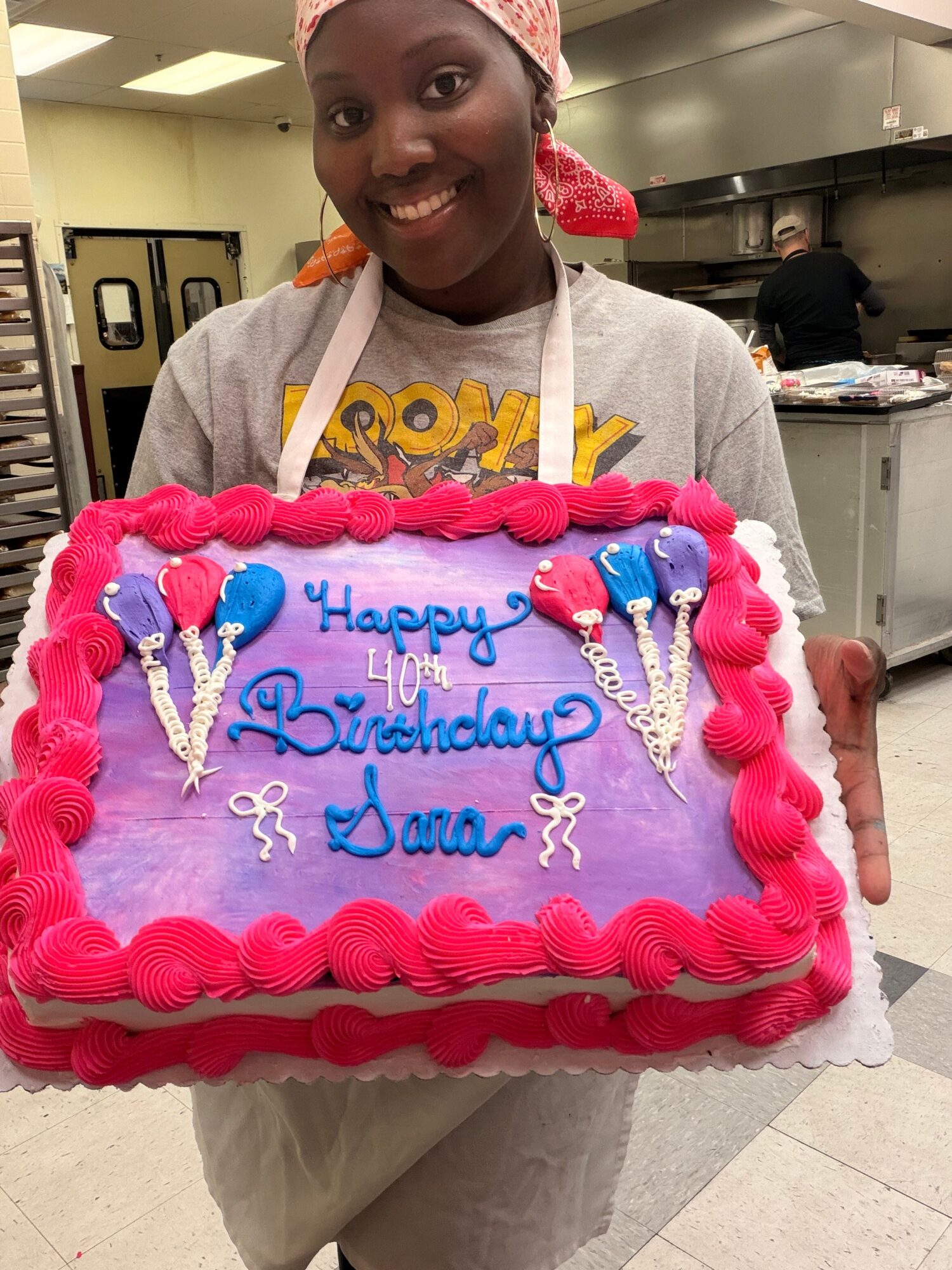 Smiling person holding a decorated birthday cake with pink, purple, and blue icing, celebrating a 40th birthday.