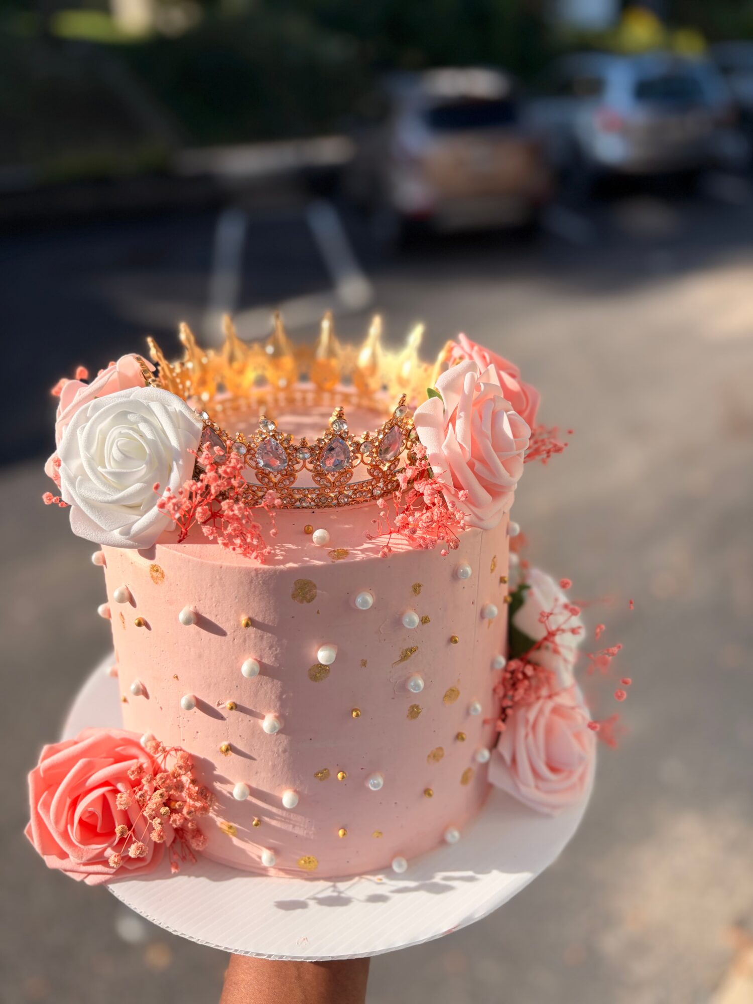 Pink birthday cake with roses, gold dots, and a crown on top, on a white plate outdoors.