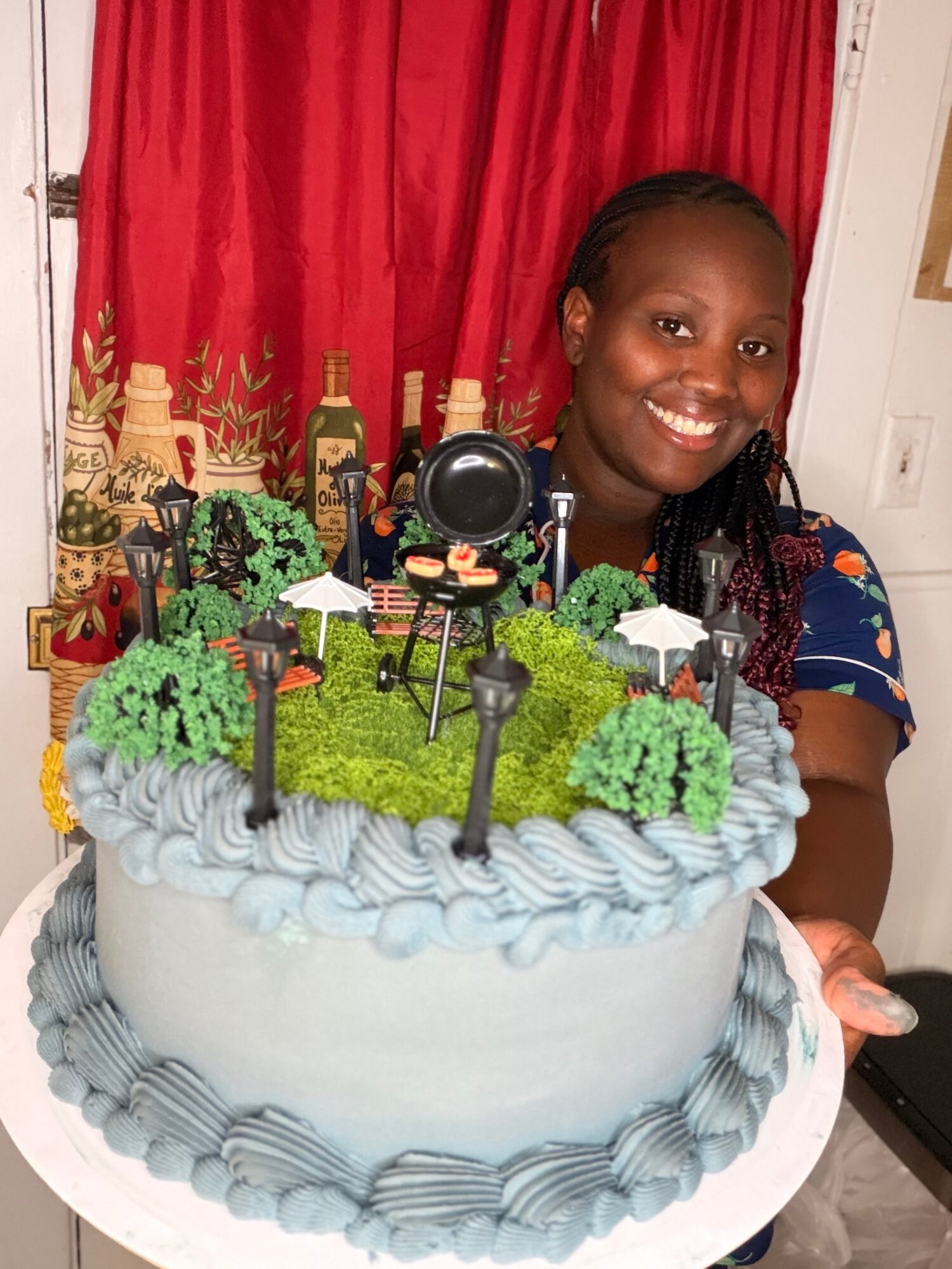 Smiling woman holding a decorated birthday cake with a camera on top, red curtains in background.