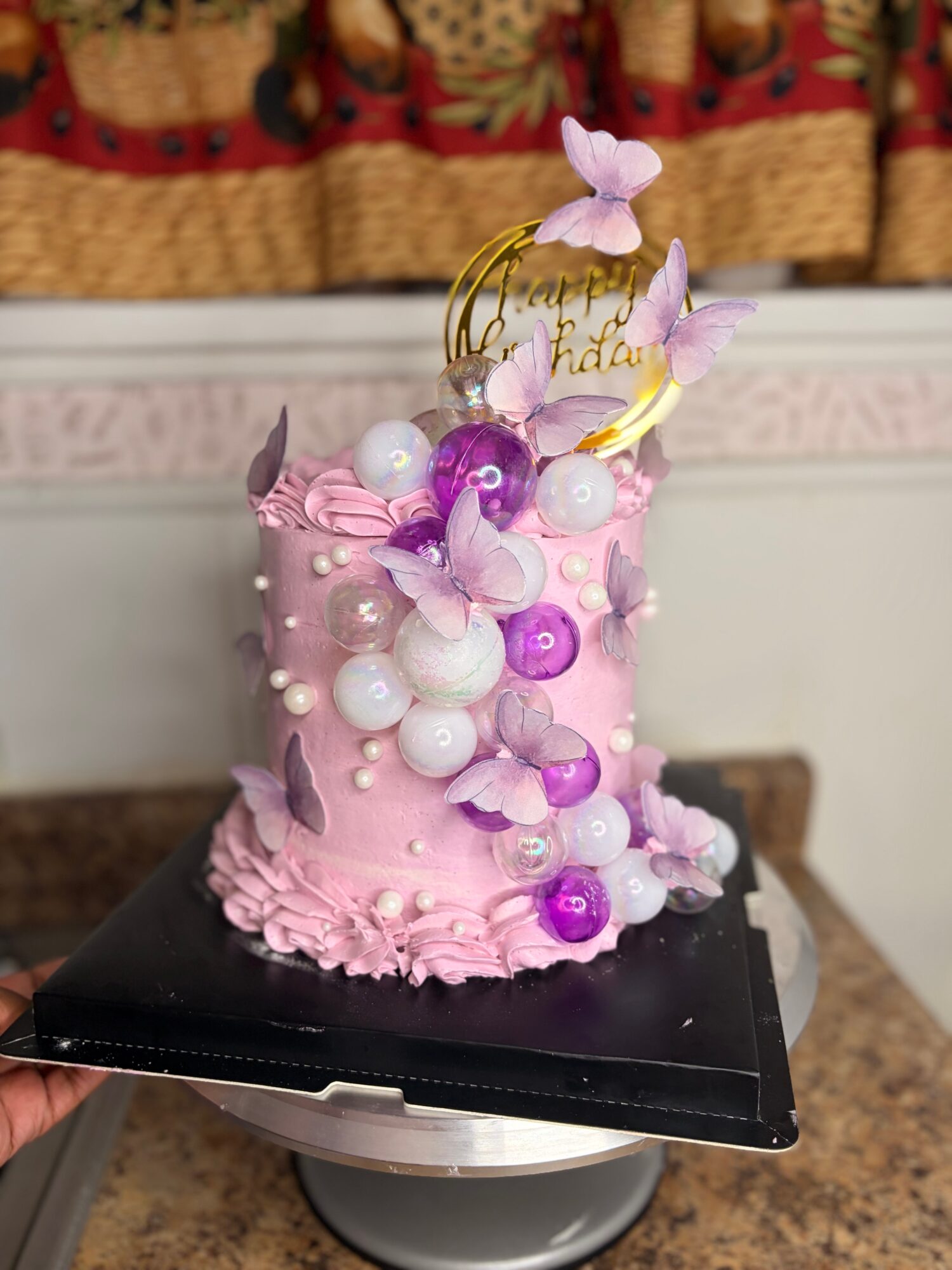 Pink birthday cake decorated with purple and white candies, butterflies, and a gold topper, on a black cake board.