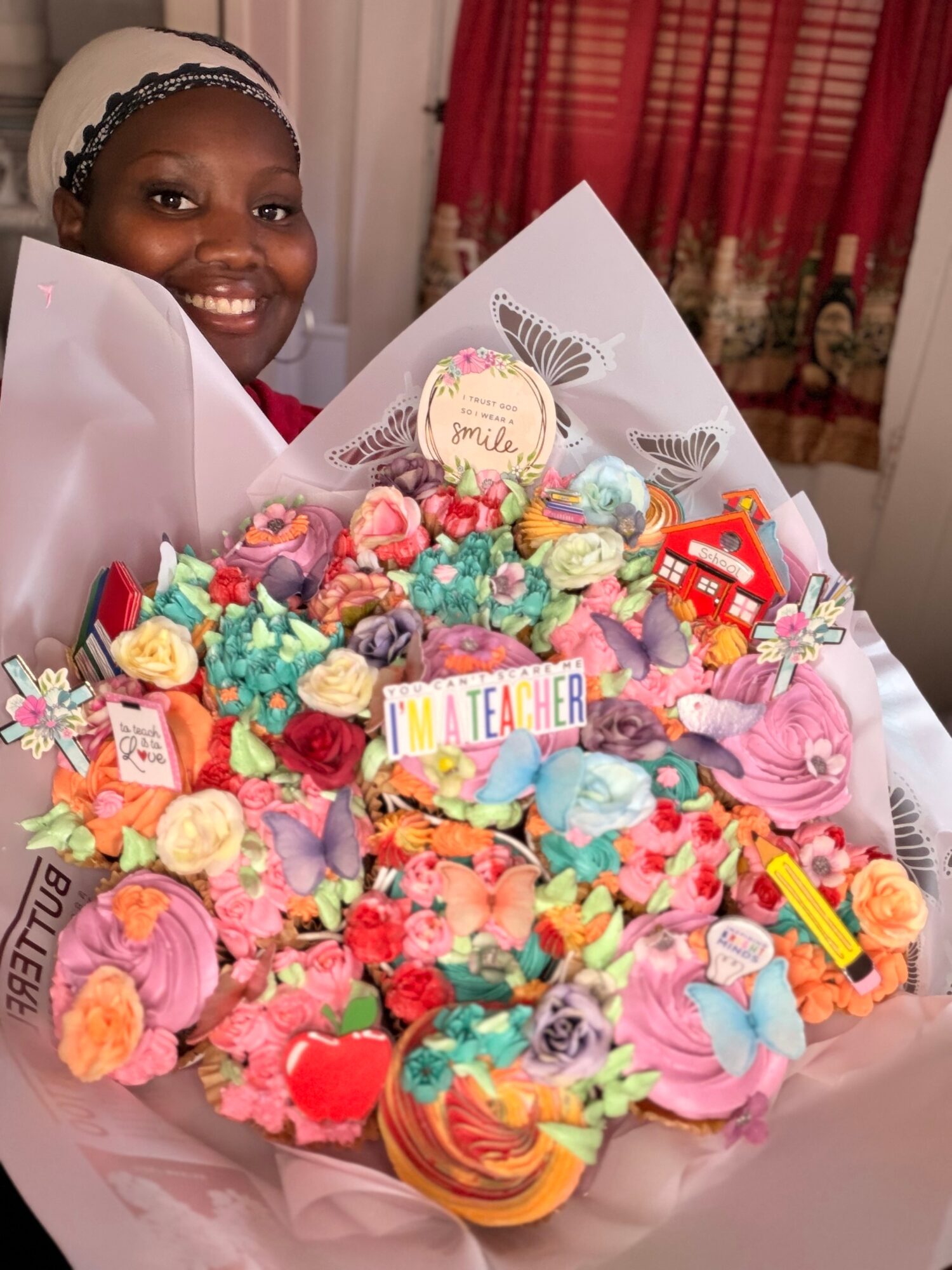 Smiling woman with a headscarf behind a large colorful bouquet of cupcakes and candies, in a room with red curtains.
