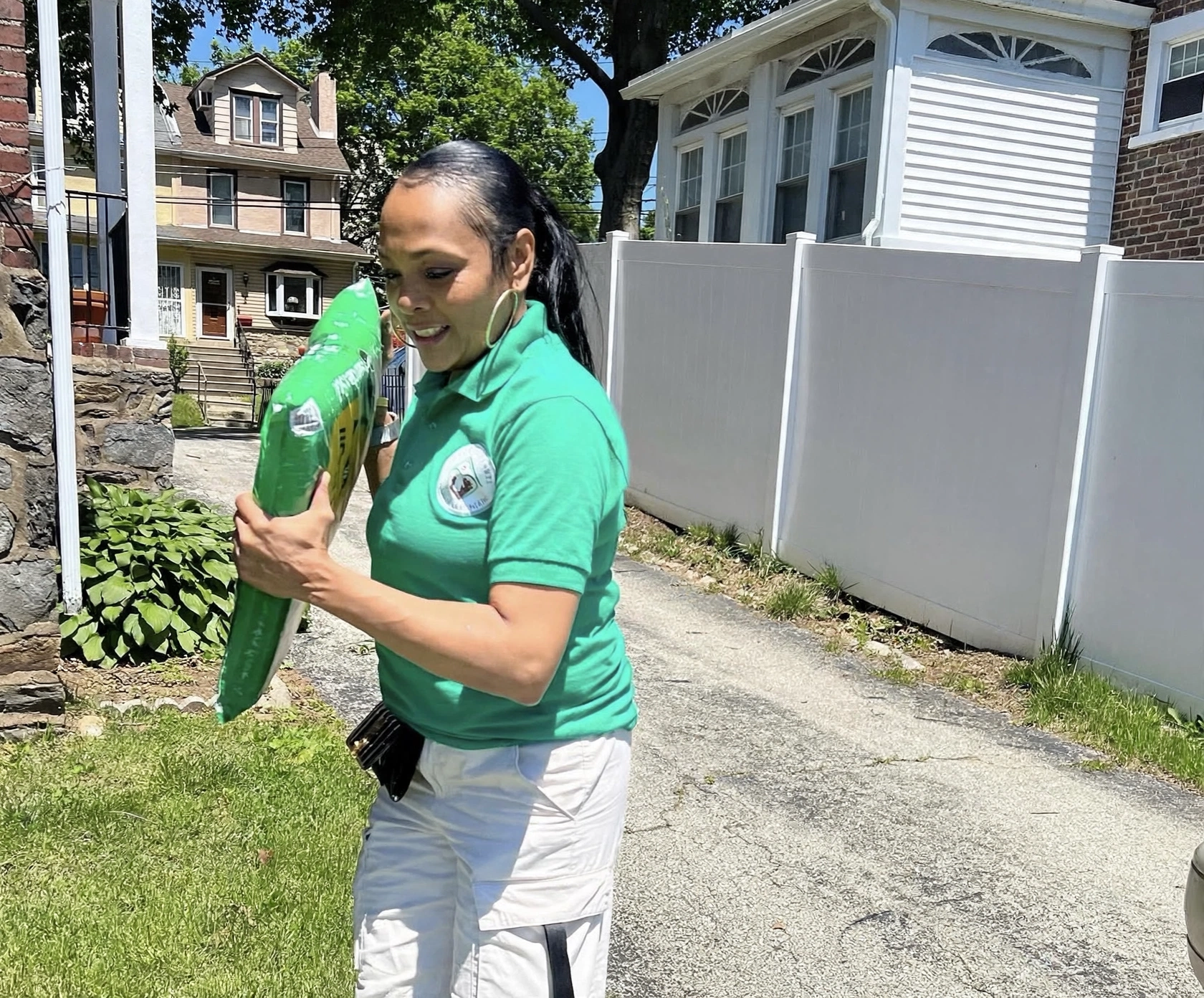 Young woman in green shirt holding a large green object outdoors, smiling, with houses and trees in background.