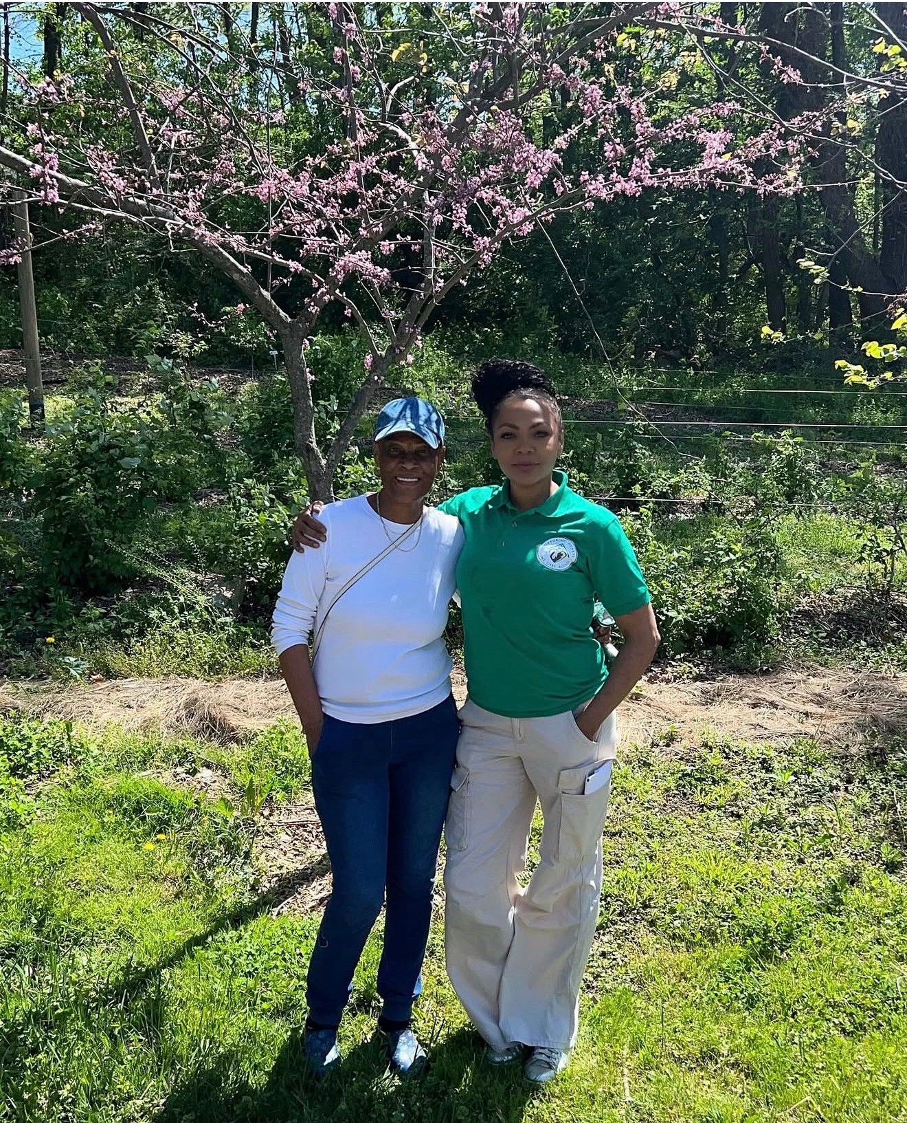 Two women standing outdoors under a pink flowering tree, smiling, with greenery and trees in the background.