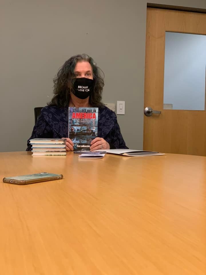 Woman with curly hair wearing a black face mask sitting at a table with books, in a room with a door and gray wall.