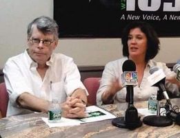 Two people sit at a table during a press conference, with microphones and papers in front of them.