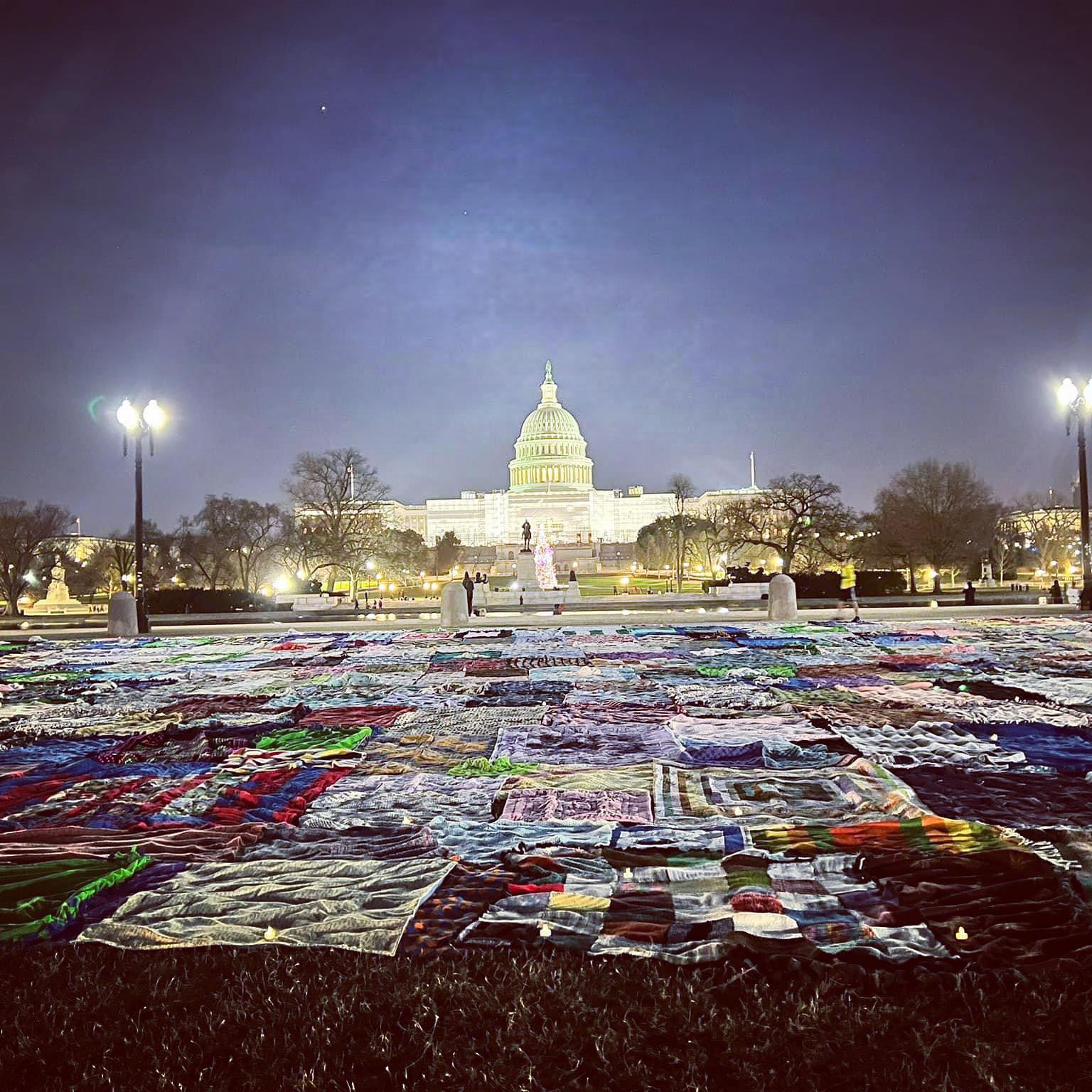 Nighttime view of the Capitol building with colorful textiles spread on the ground in front, illuminated by streetlights.