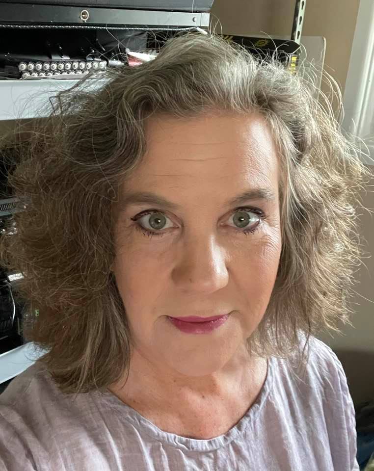 Portrait of a woman with curly hair, wearing a light-colored top, looking directly at the camera indoors.
