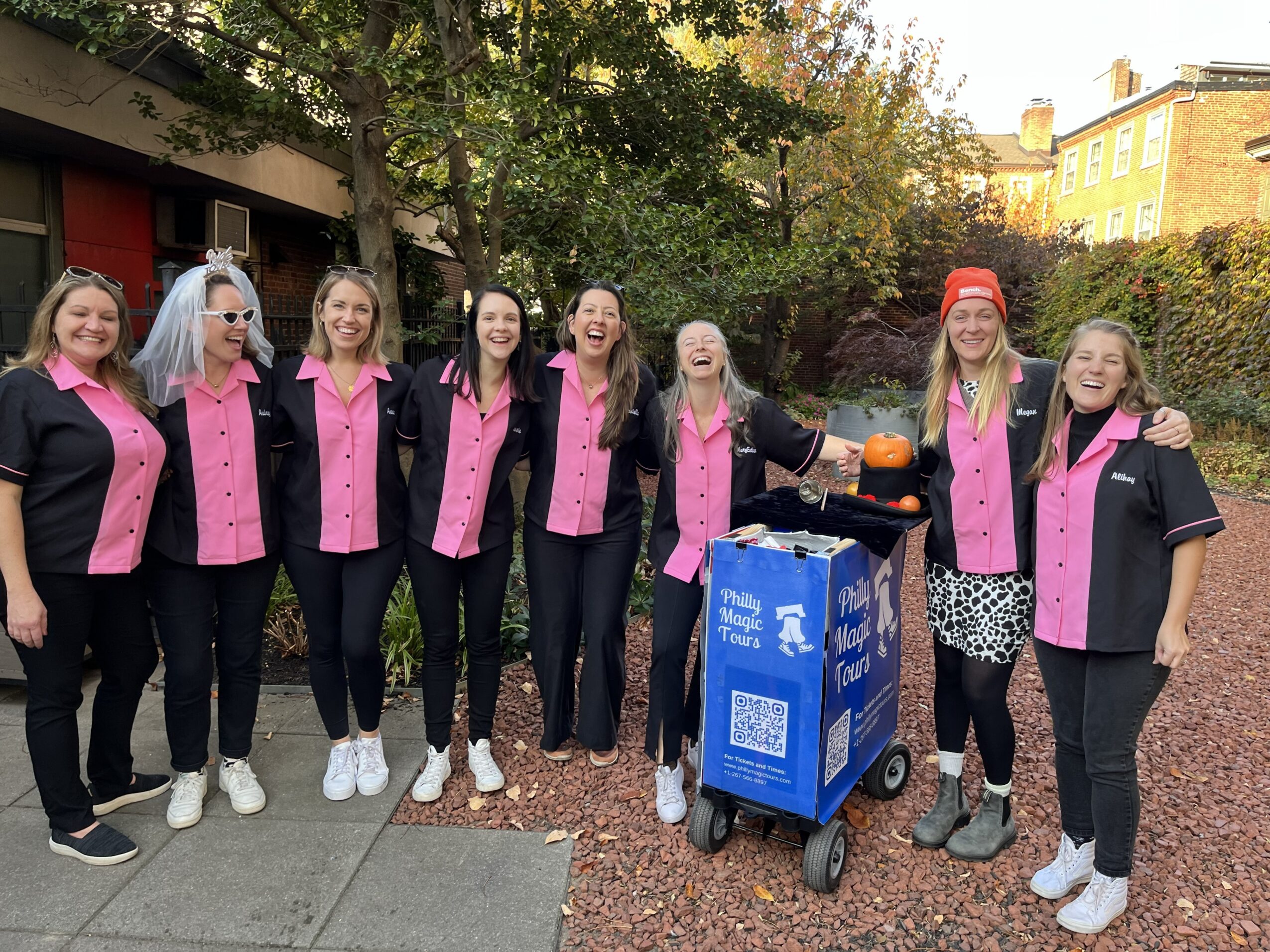 Group of eight women standing outdoors, smiling, wearing matching pink and black jackets, with a blue cart and trees in background.
