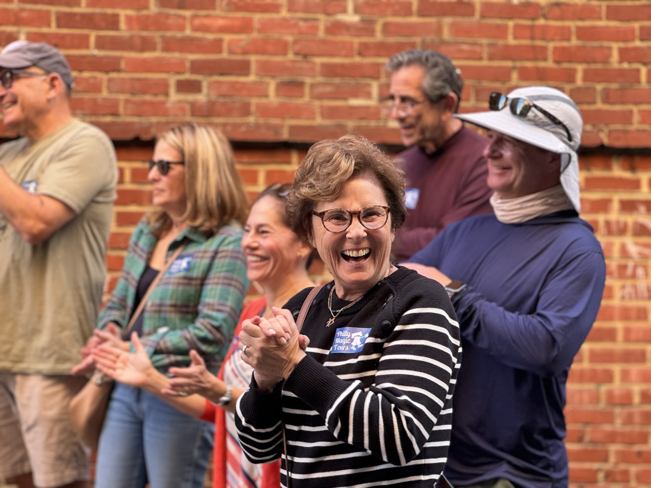 Group of six people smiling and clapping outdoors against a brick wall, some wearing sunglasses and hats.
