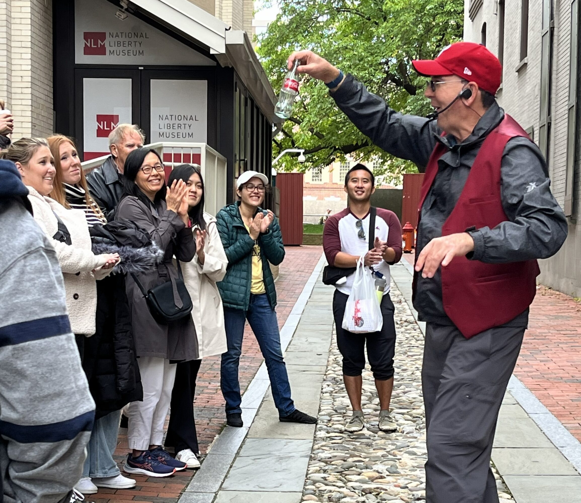 A man in a red cap and vest performs a street magic trick for a group of onlookers outside a museum.