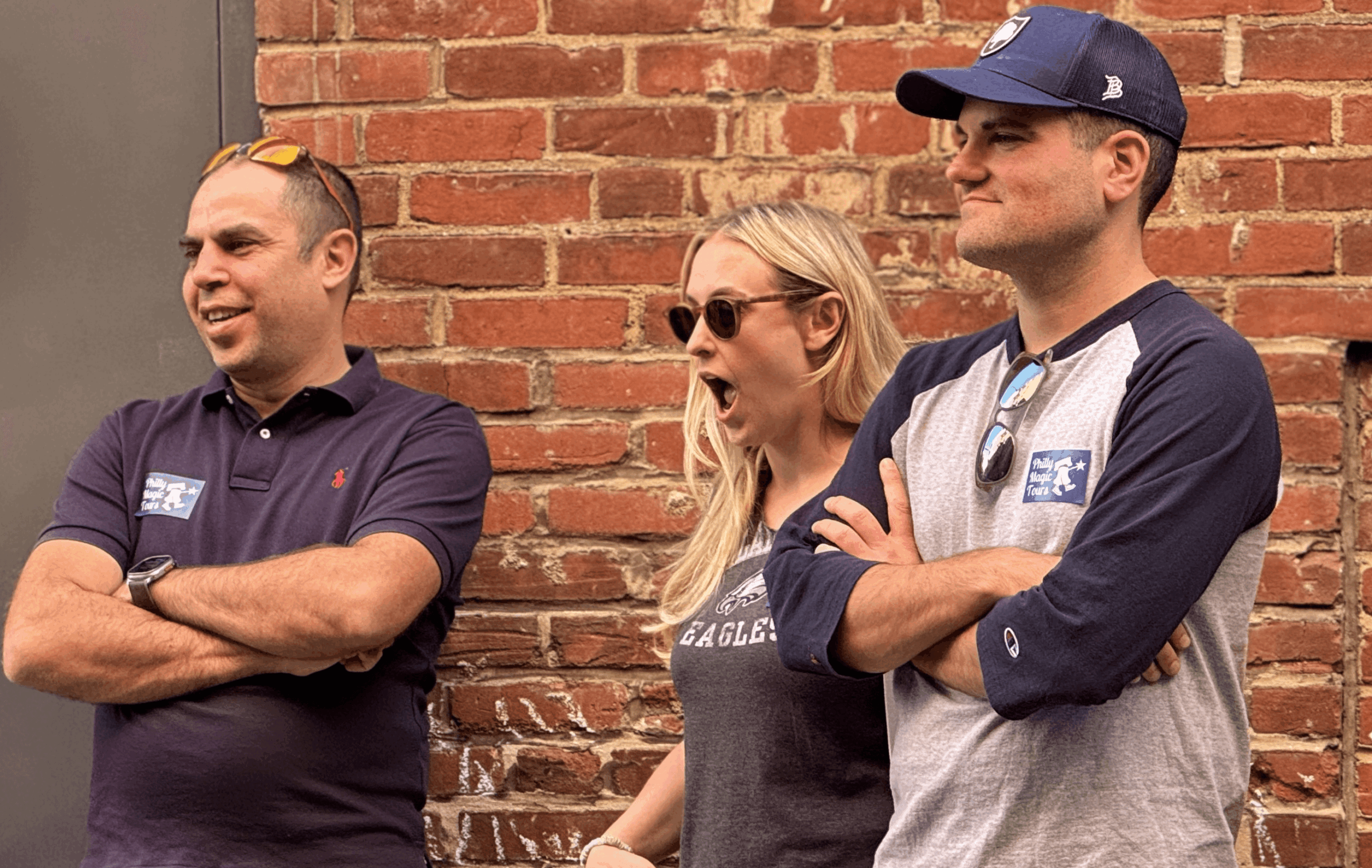 Three people standing outdoors in front of a brick wall, smiling and talking, with one woman wearing sunglasses.