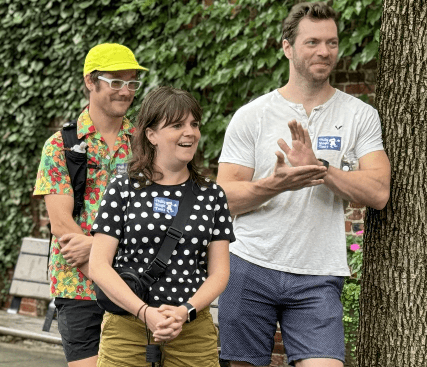 Three people standing outdoors near a tree, smiling and clapping, with greenery in the background.
