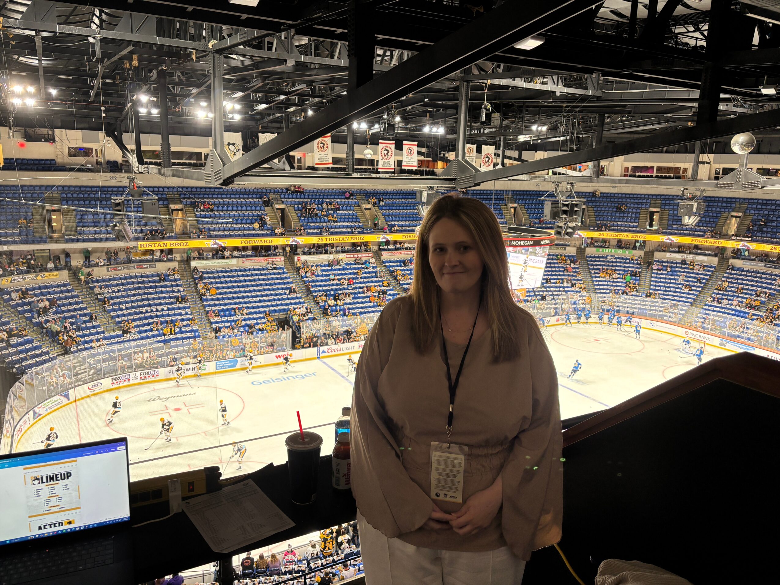Woman standing in front of a hockey arena with players on the ice, seating, and arena lights visible.