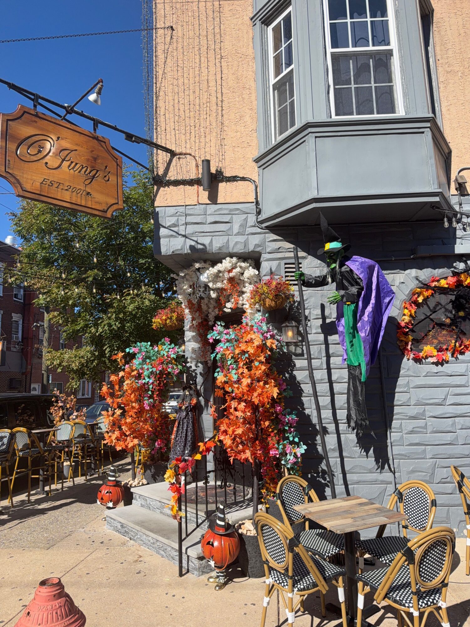 Outdoor scene with chairs, a sign, and colorful autumn decorations on a patio, with a building in the background.